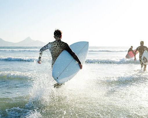Three teenage boys wearing their wetsuits carrying their surfboards run into the sea on a sunny Summers day.